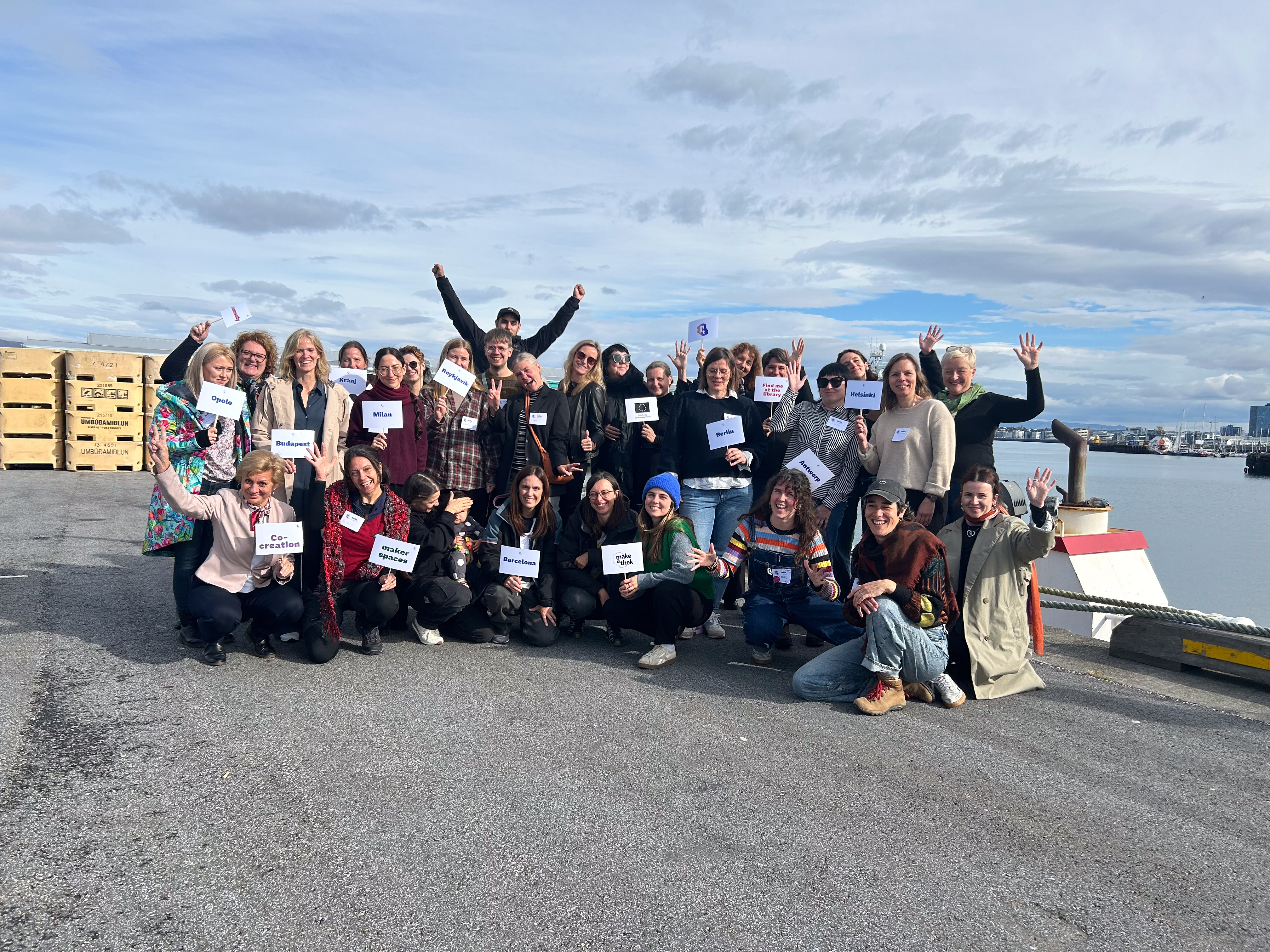 A group of participants, project partners, and librarians gathered on a pier for a consortium meeting, holding signs representing cities and organizations, as they wave to the camera.